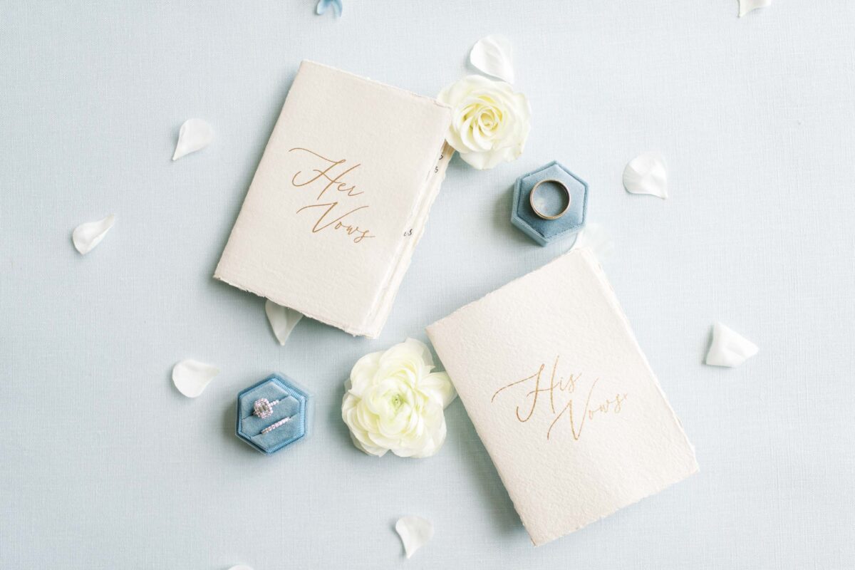 his and her vows next to white flowers and the wedding bands in blue velvet boxes