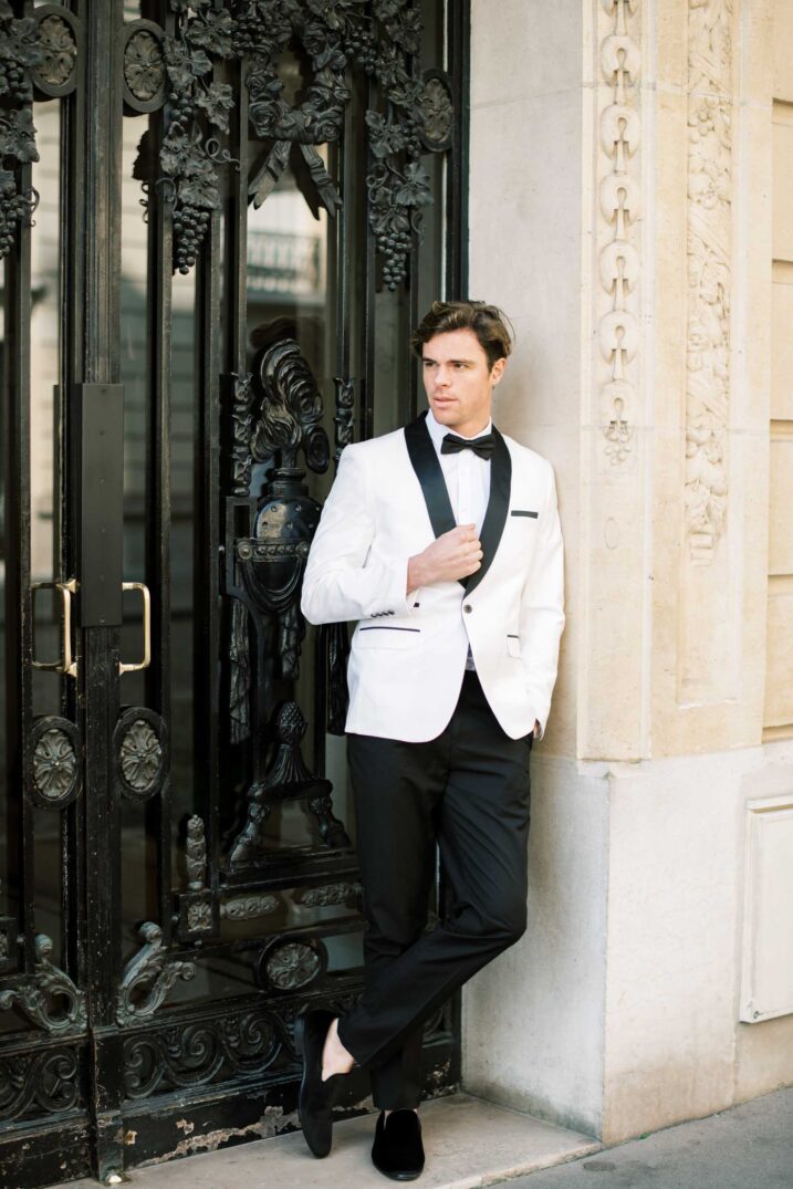 groom wearing a white tuxedo standing against a black metal door in paris 