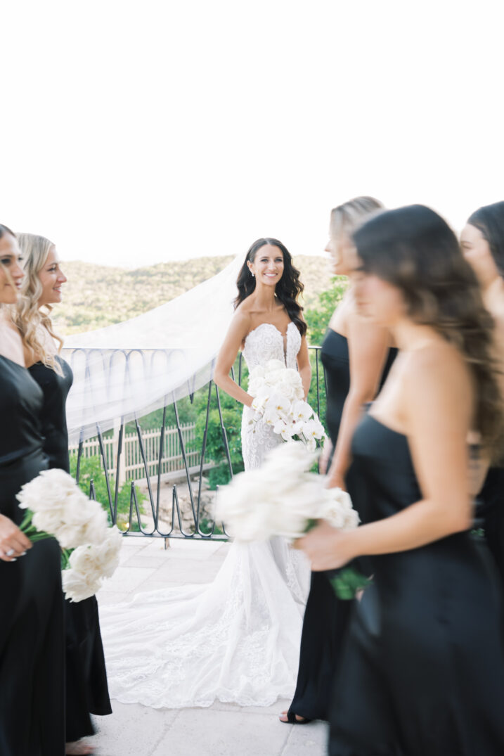 smiling bride wedding dress veil in wind bridesmaids walking by white florals 